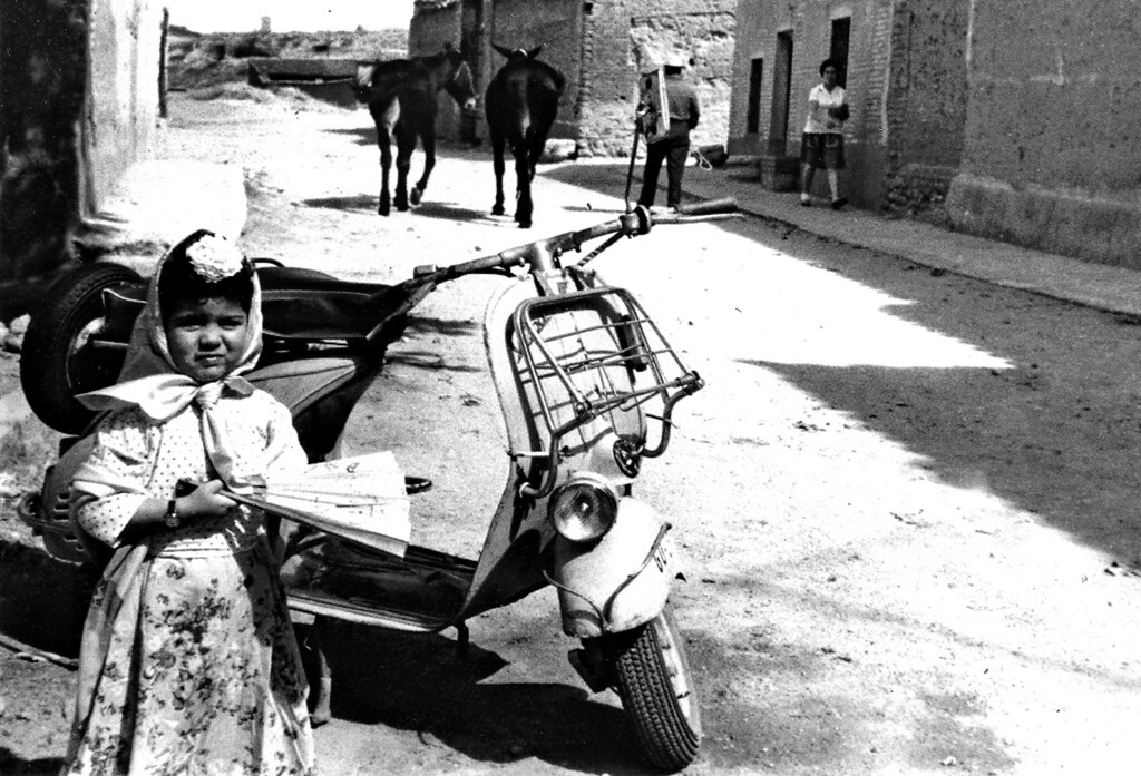 Una niña posa delante de una Vespa en un pueblo con dos burros al fondo