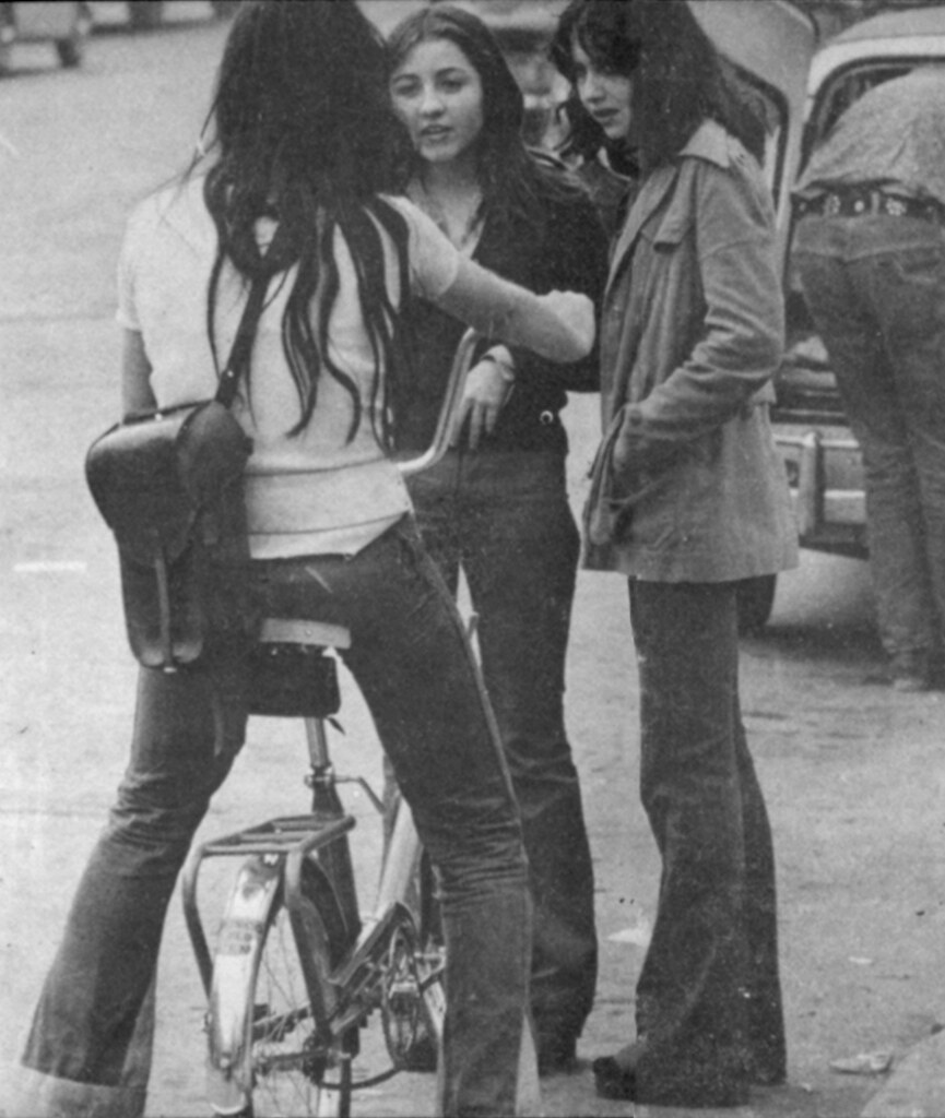 Foto en blanco y negro de tres mujeres jóvenes con ropa de los 70, una de ellas en bicicleta