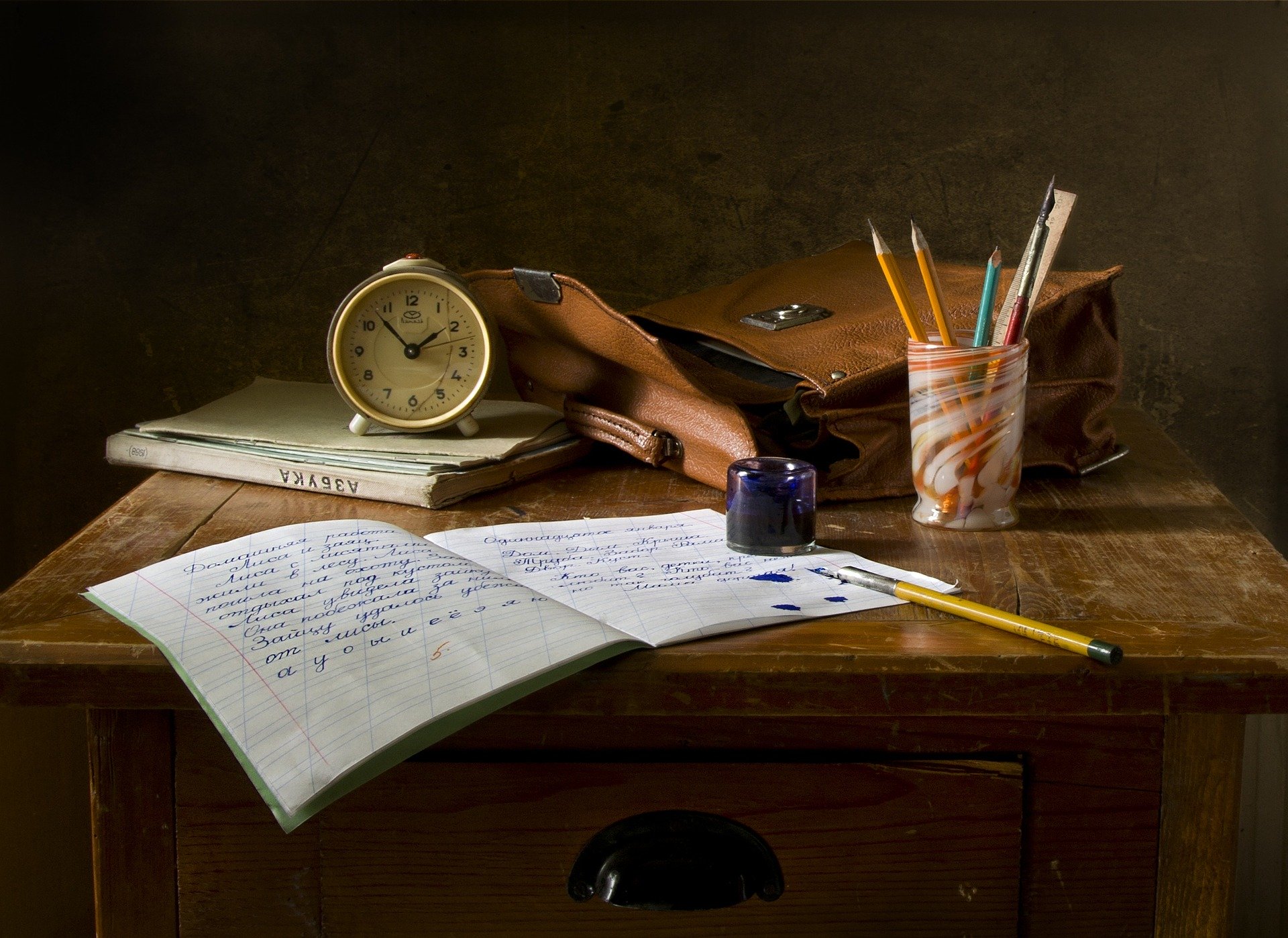 Un bodegón compuesto por una libreta, una pluma con su bote de tinta, una cartera escolar abierta y un reloj analógico clásico.