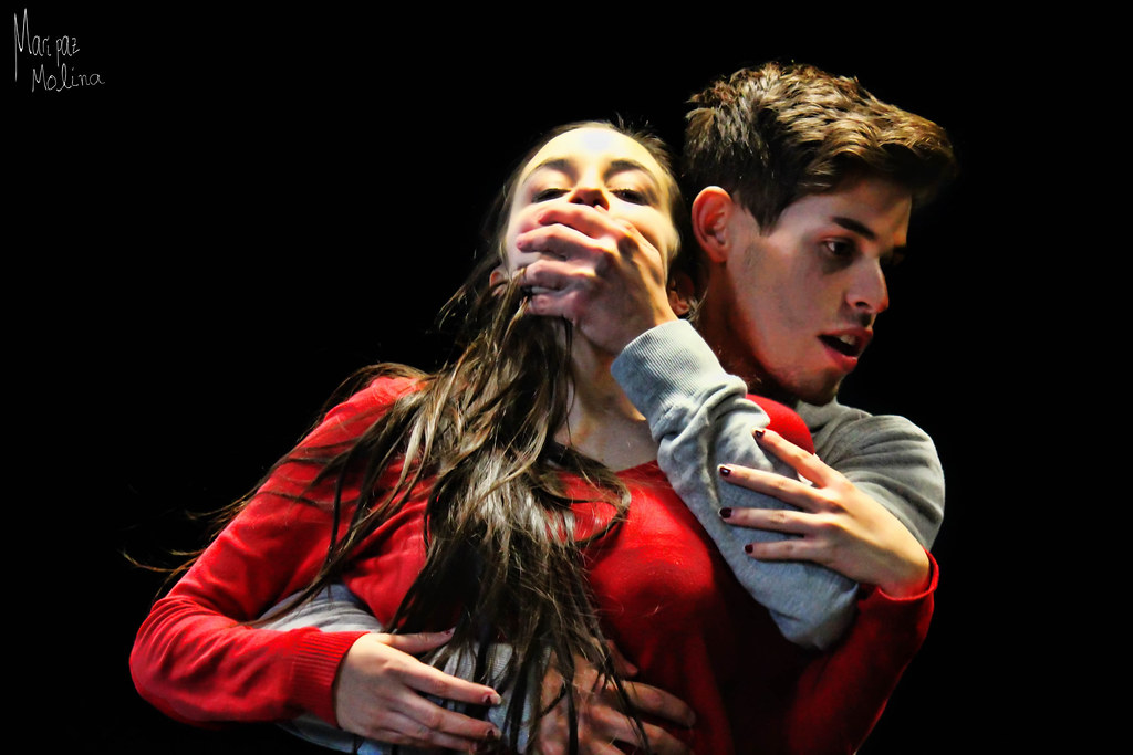 Chico agarra a una chica por detrás y le tapa la boca. La chica lleva un vestido rojo. 
