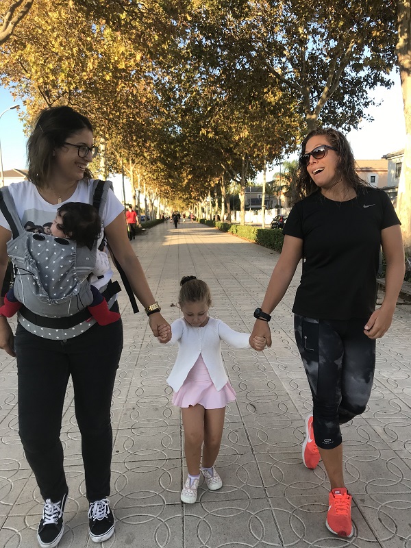Una familia formada por dos madres, un bebé en la mochila de una de ellas y una niña pequeña charlando mientras dan un paseo.