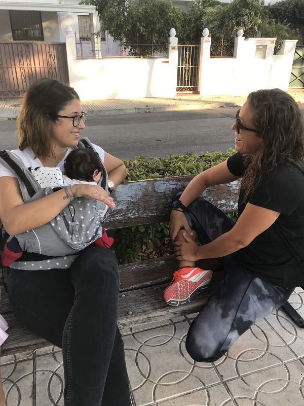 Dos chicas, una de ellas con un bebé en la mochila, charlando en un banco
