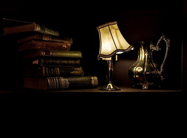 Bodegón lámpara luz Libros amontonados en una mesa cerca de una jarra de metal e iluminados por una lámpara torcida.